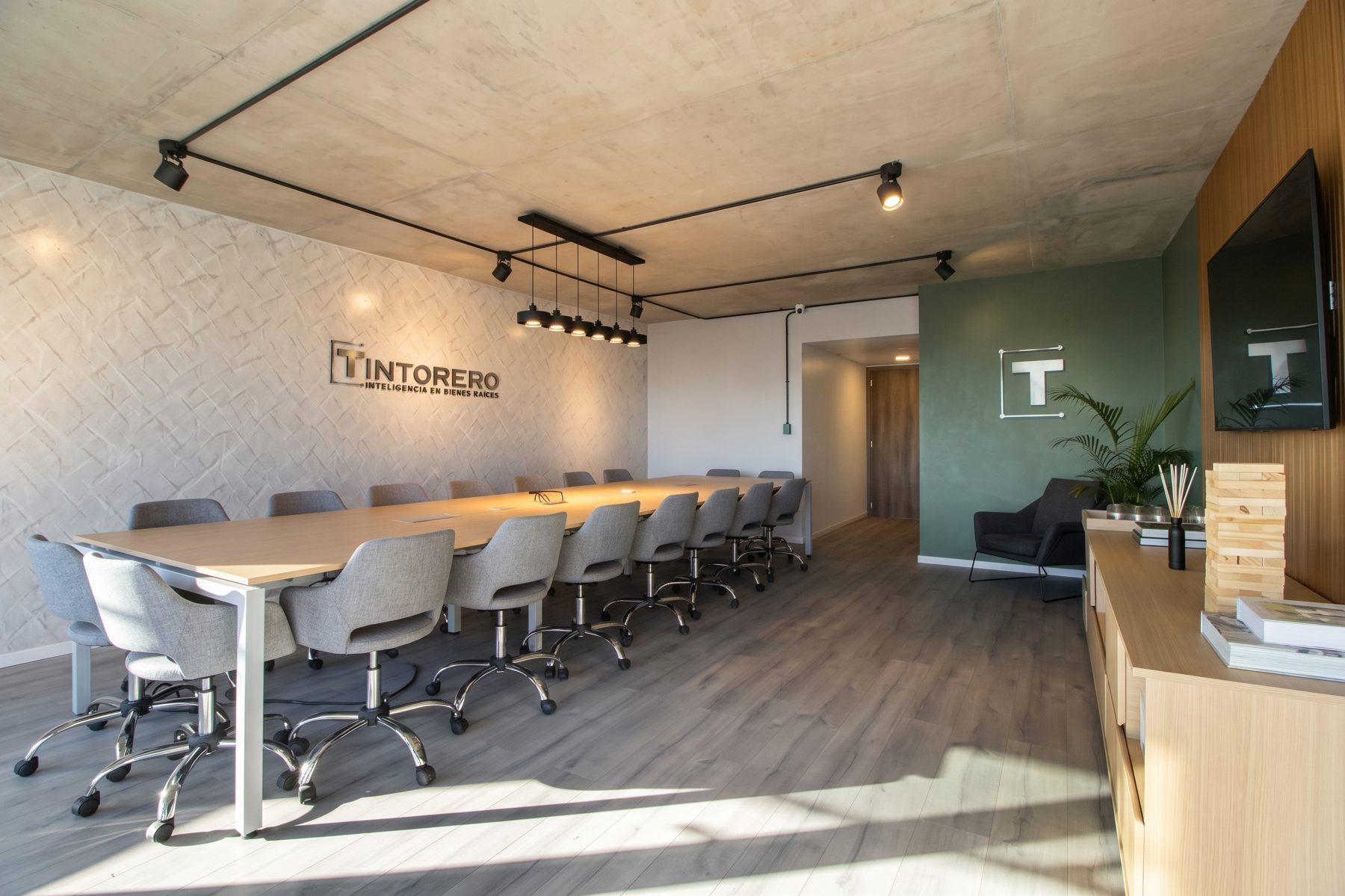 Sala de reuniones de Tintorero con mesa larga para 18 personas, sillas grises, pared verde con el logo y muro texturado